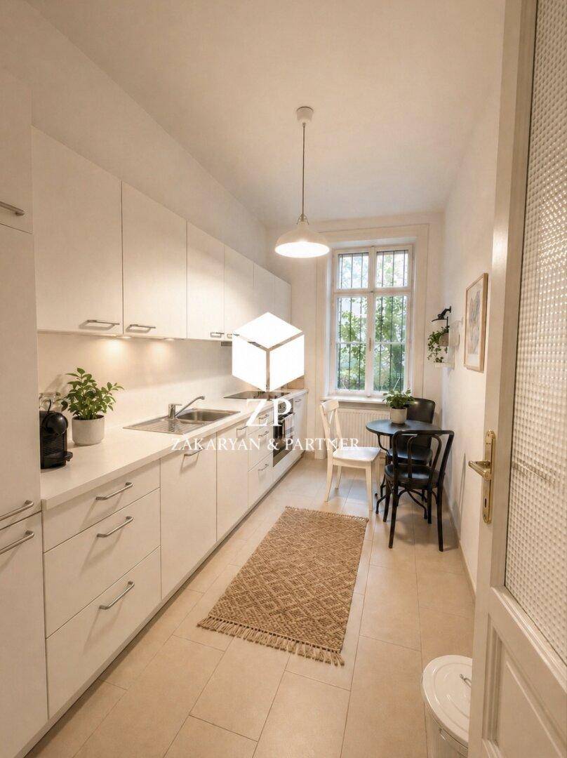 Schmale, helle Küche mit weißen Schränken, Fliesenboden und kleinem Essbereich am Fenster.