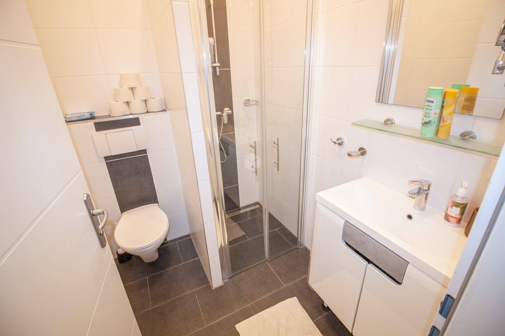 Kleines, modernes Badezimmer mit Duschkabine, wandhängendem WC und Waschbecken, in Weiß und Grau gefliest.