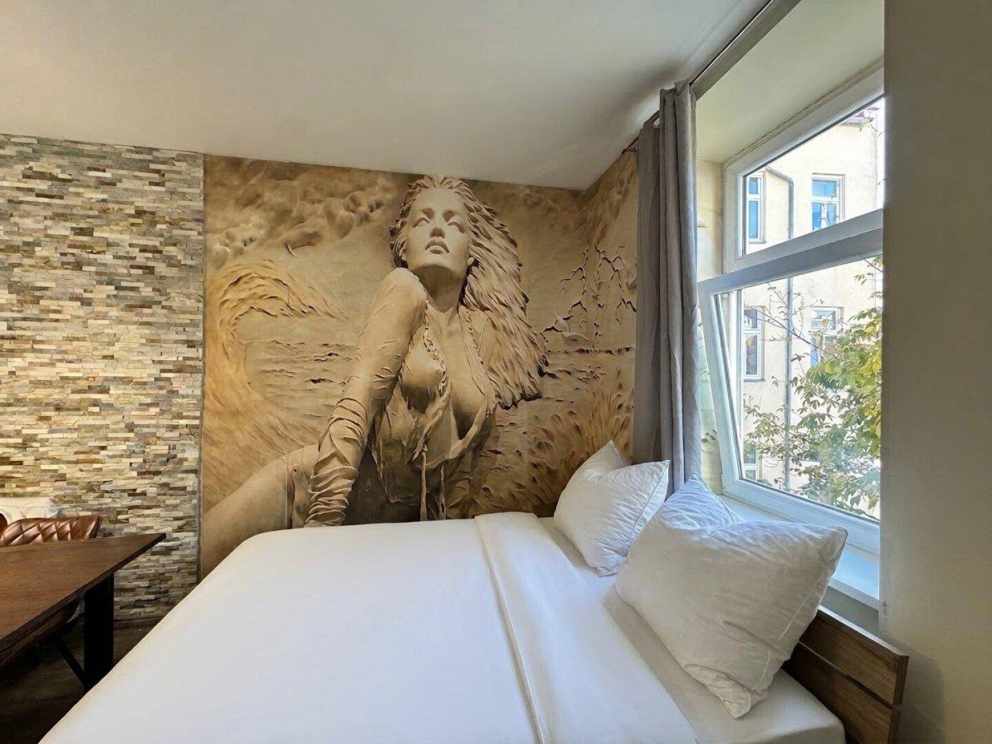 Schlafzimmer mit Doppelbett am Fenster und einer beeindruckenden Wandmalerei, die eine Frau darstellt.