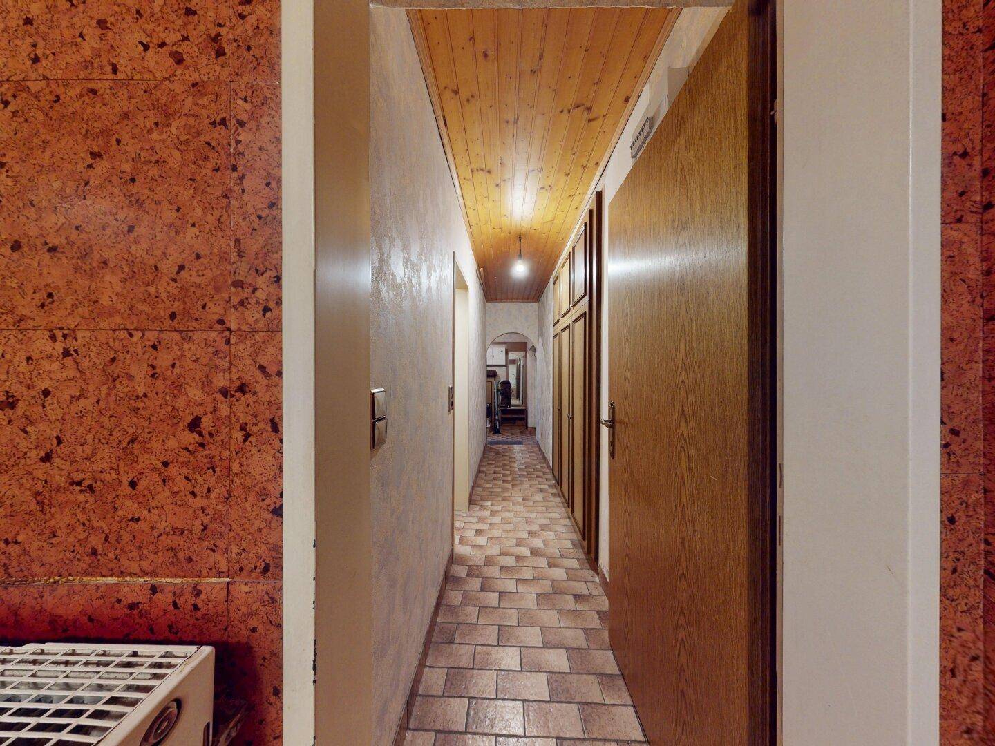 Langer Flur mit gefliestem Boden, Holzdecke und Holztüren, der zu weiteren Räumen führt.