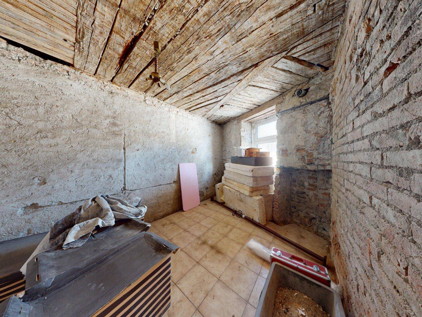 Dachbodenraum im Rohzustand mit freigelegten Ziegelwänden, Holzbalkendecke und einem Fenster.