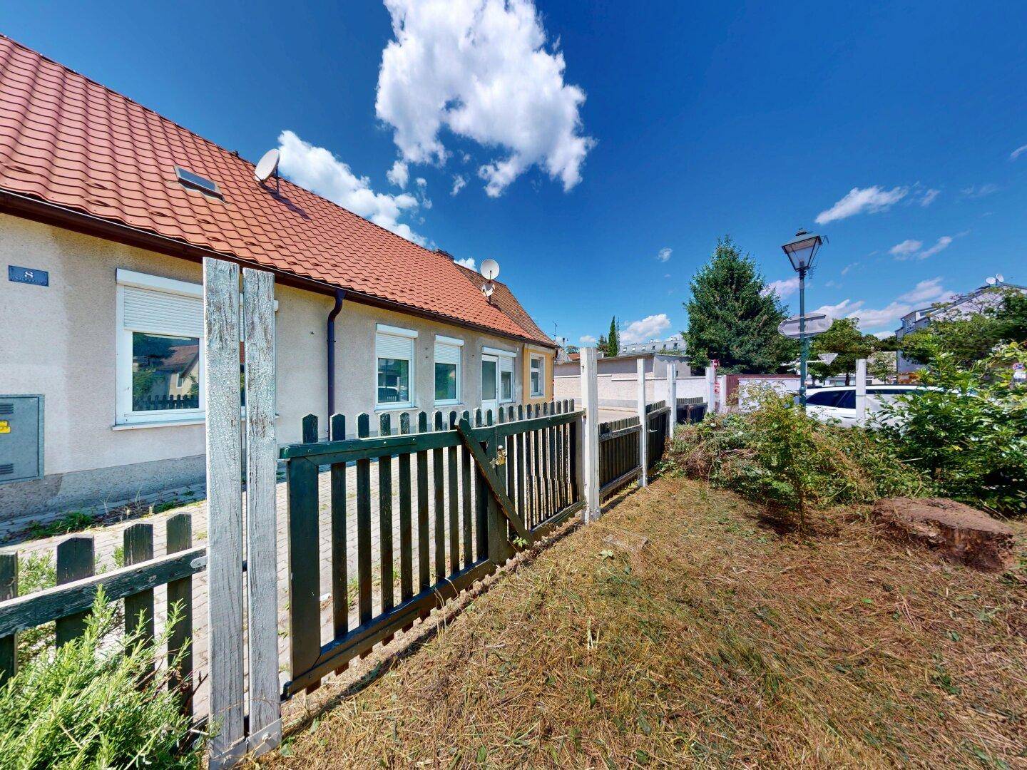 Seitenansicht des Hauses mit Holzzaun und einem naturbelassenen Gartenbereich unter blauem Himmel.