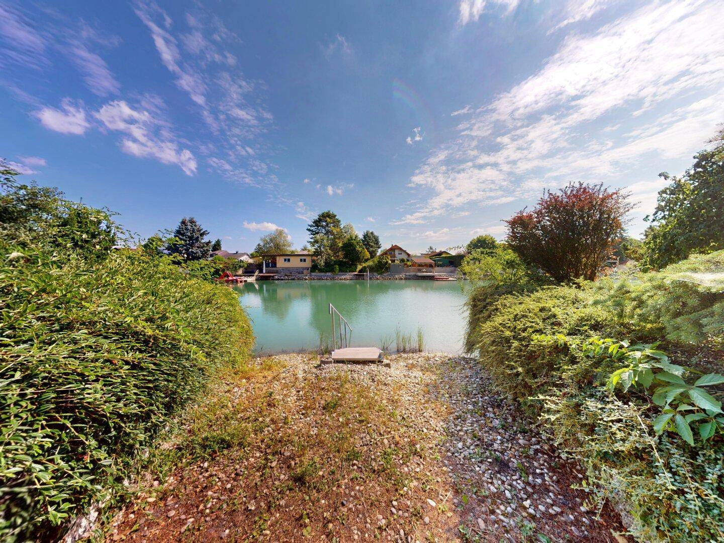 Panoramablick auf den See mit Häusern am gegenüberliegenden Ufer und üppiger Ufervegetation.