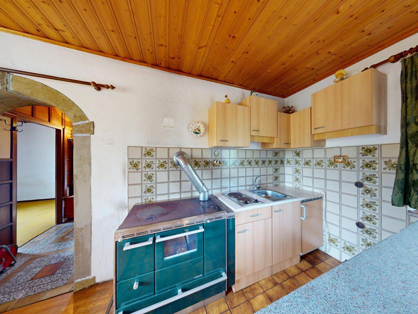 Küchenbereich mit einem markanten grünen Holzofen, Spüle und hellen Holzschränken.