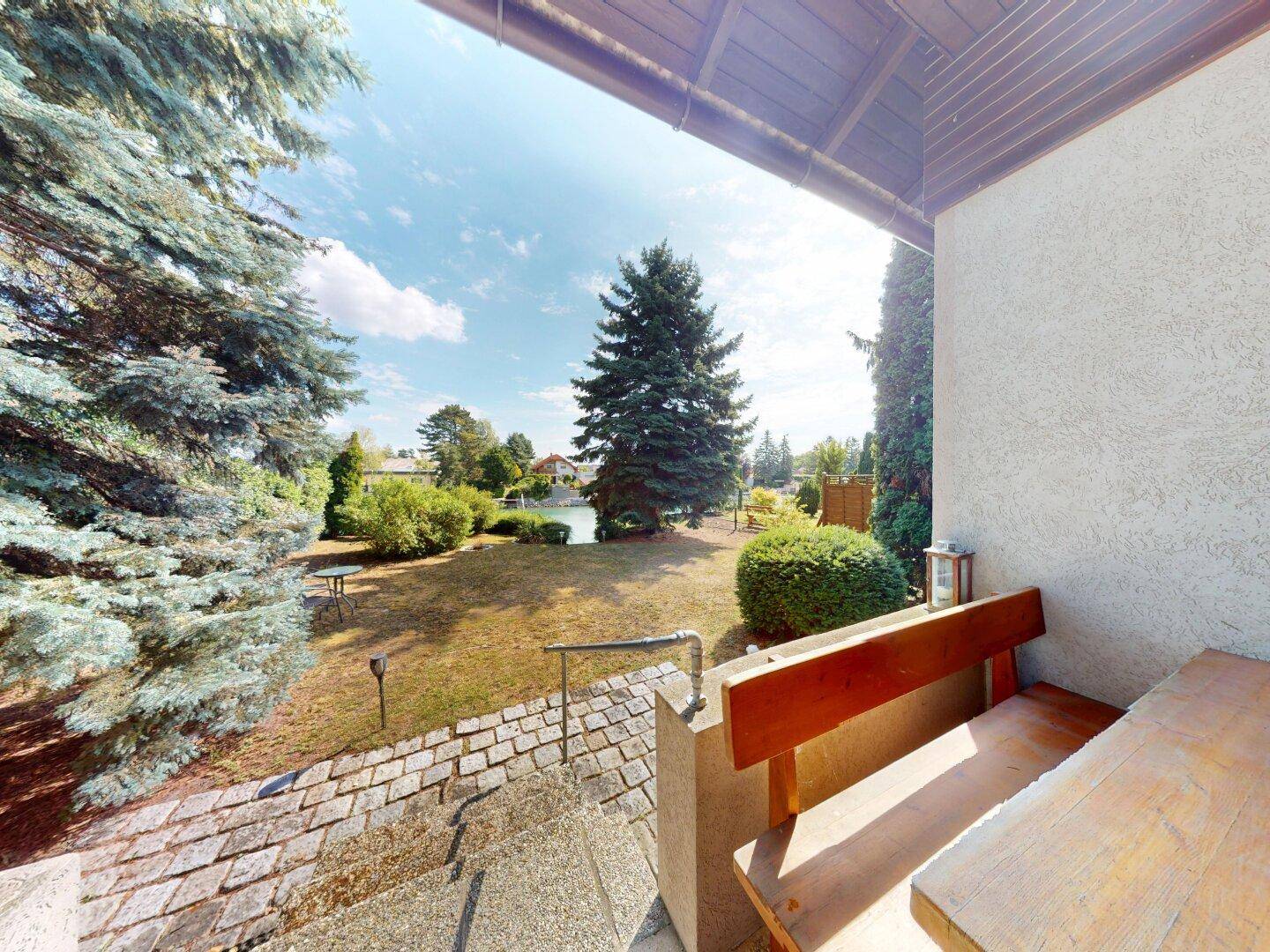 Überdachte Terrasse mit Holzmöbeln und Treppe zum Garten, umgeben von viel Grün.