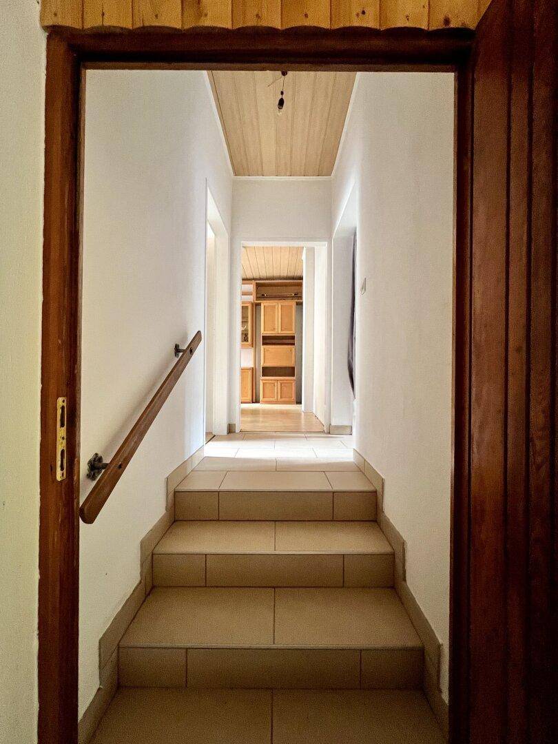 Gefliester Treppenaufgang mit Holzgeländer führt zu einem Flur mit Holzdecke.