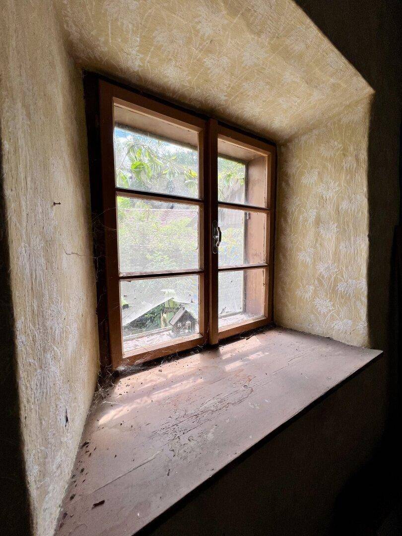 Kleines Holzfenster mit Blick ins Grüne, umgeben von alter, gemusterter Tapete.