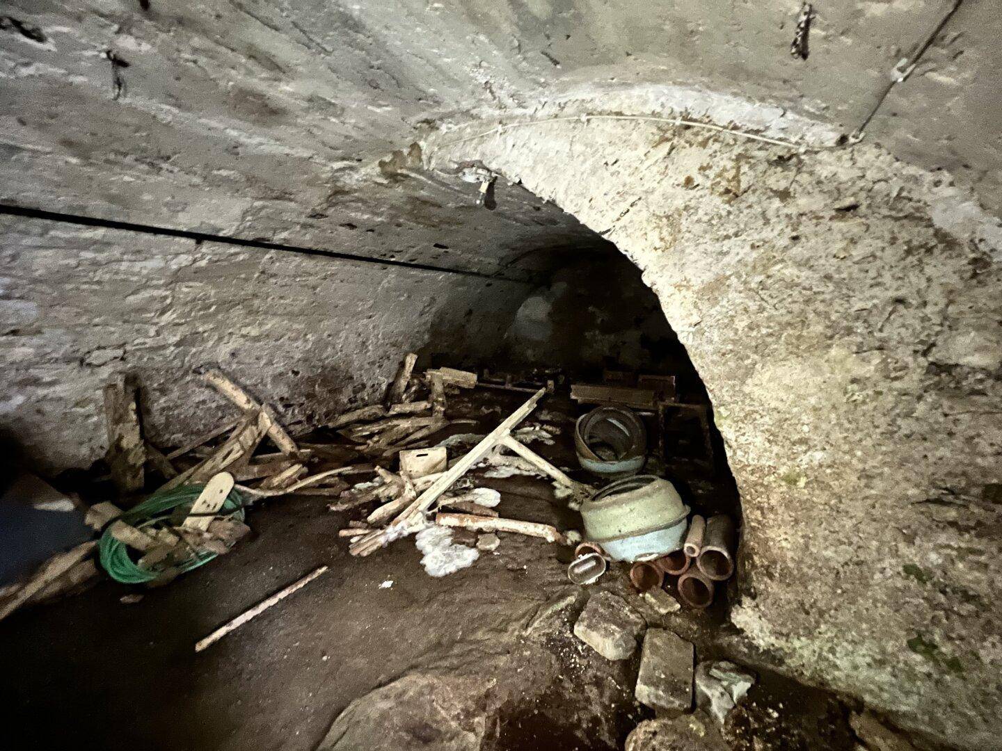 Unaufgeräumter Kellergang mit Steingewölbe und verschiedenen Materialien auf dem Boden.