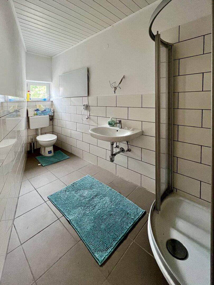 Badezimmer mit Duschkabine, Waschbecken, WC und Fenster, gefliest in Weiß und Grau.