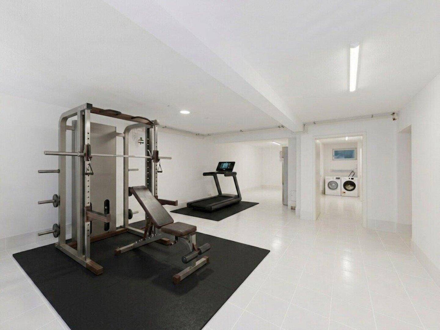 Geräumiger Fitnessraum im Untergeschoss mit verschiedenen Trainingsgeräten und Waschküche im Hintergrund.