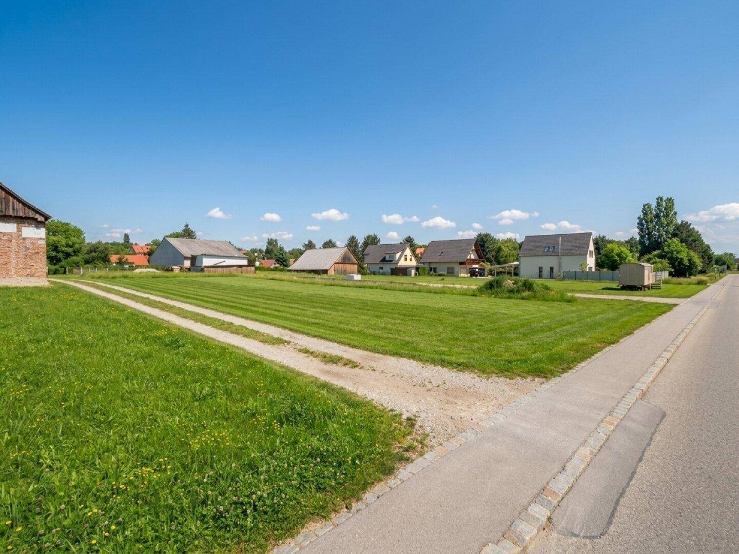 Weitläufiges grünes Grundstück mit Zufahrtsweg und Wohnhäusern im Hintergrund unter blauem Himmel.