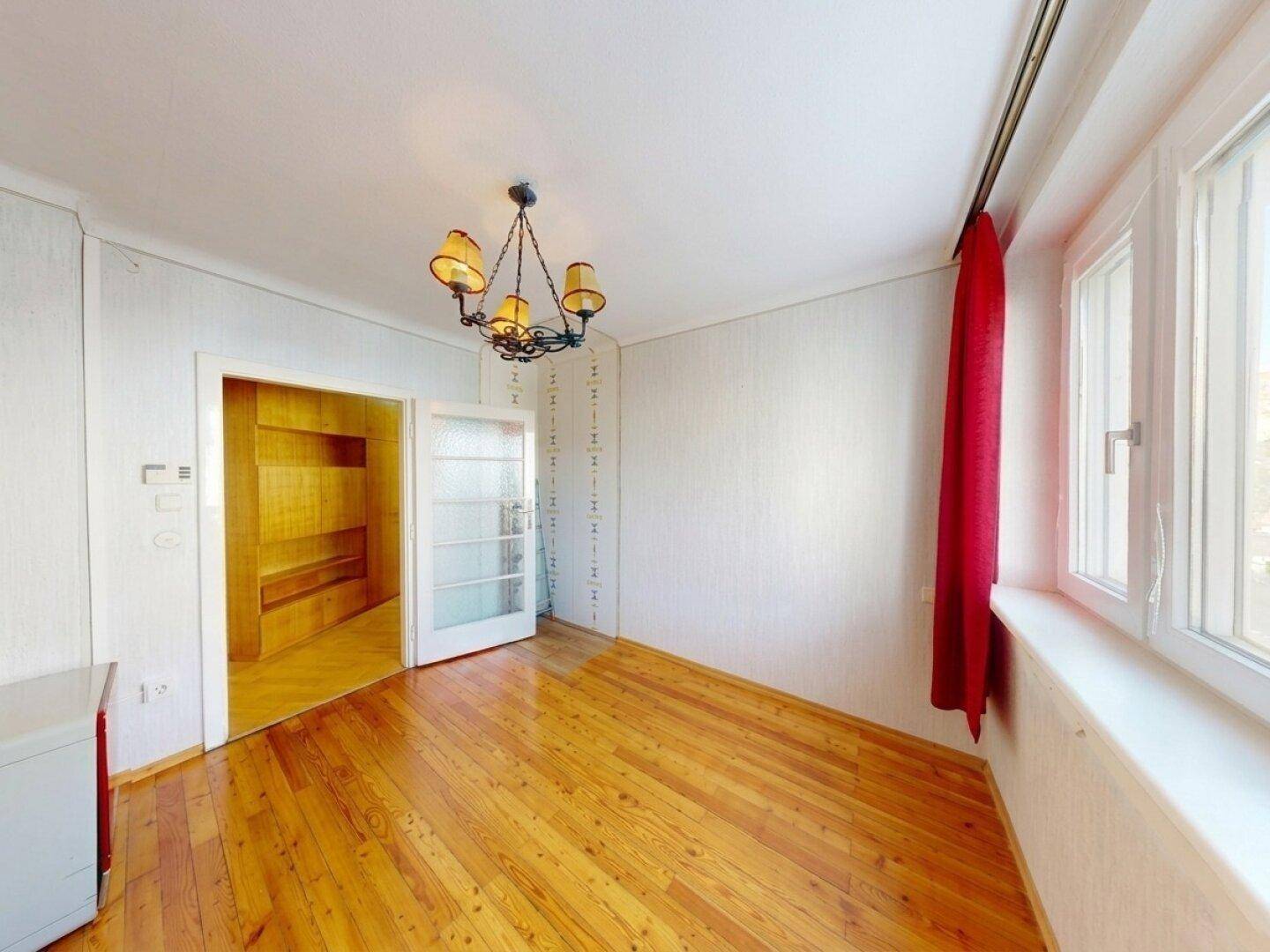 Heller Raum mit Holzboden, großem Fenster und Zugang zu einem weiteren Zimmer.