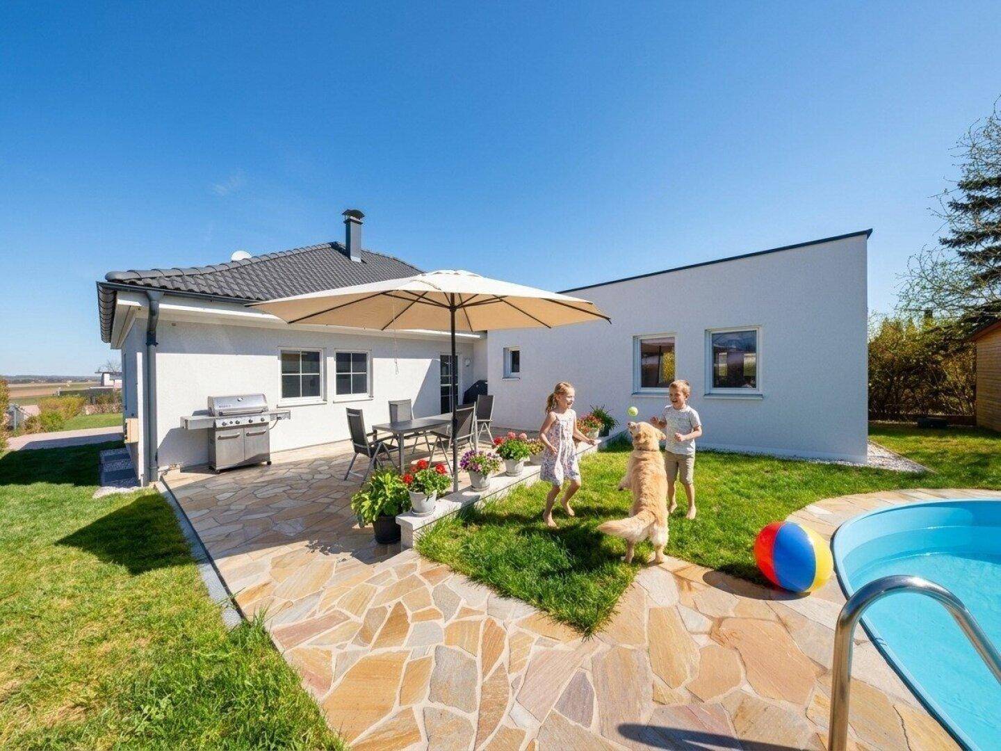 Sonniger Gartenbereich mit Terrasse, Grillplatz, kleinem Pool und spielenden Kindern.