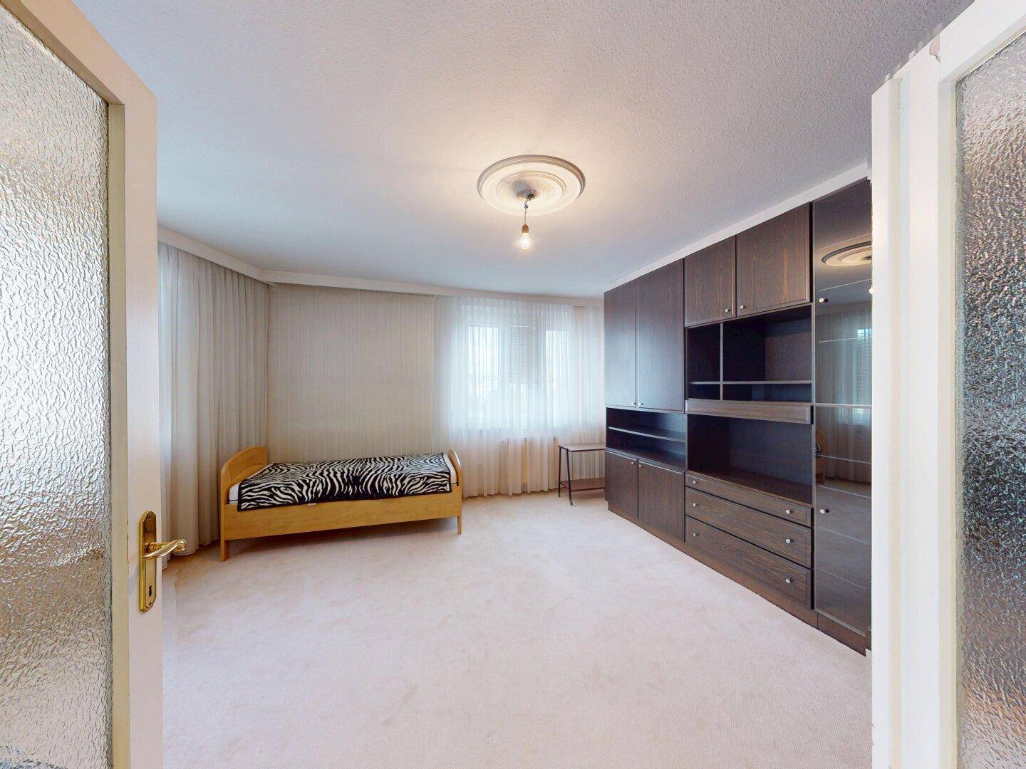 Schlafzimmer mit Einzelbett, dunklen Einbauschränken und einem großen Fenster für natürliches Licht.