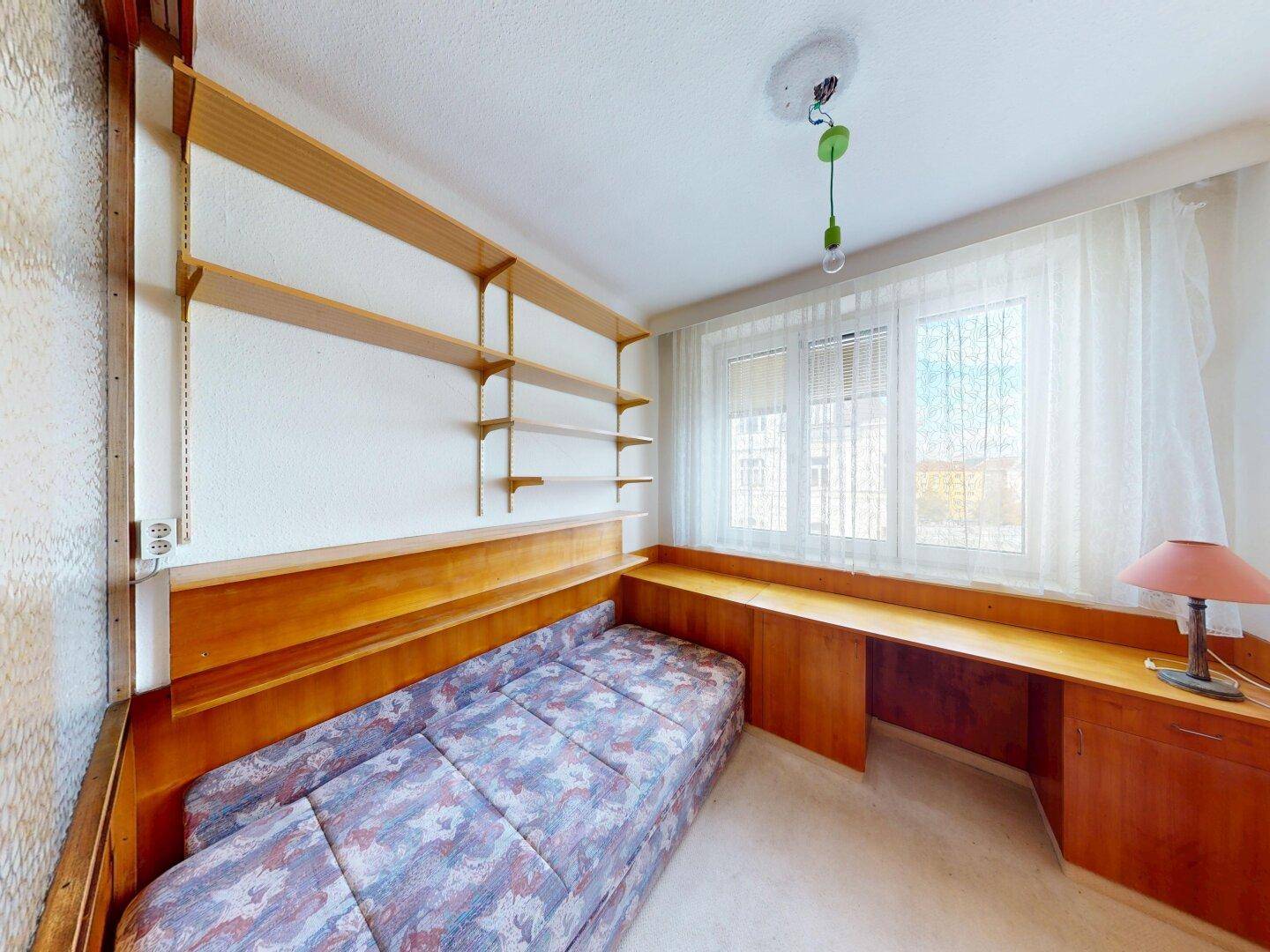 Kleines Arbeitszimmer mit integrierten Holzregalen, Schreibtisch und einem Schlafsofa am Fenster.