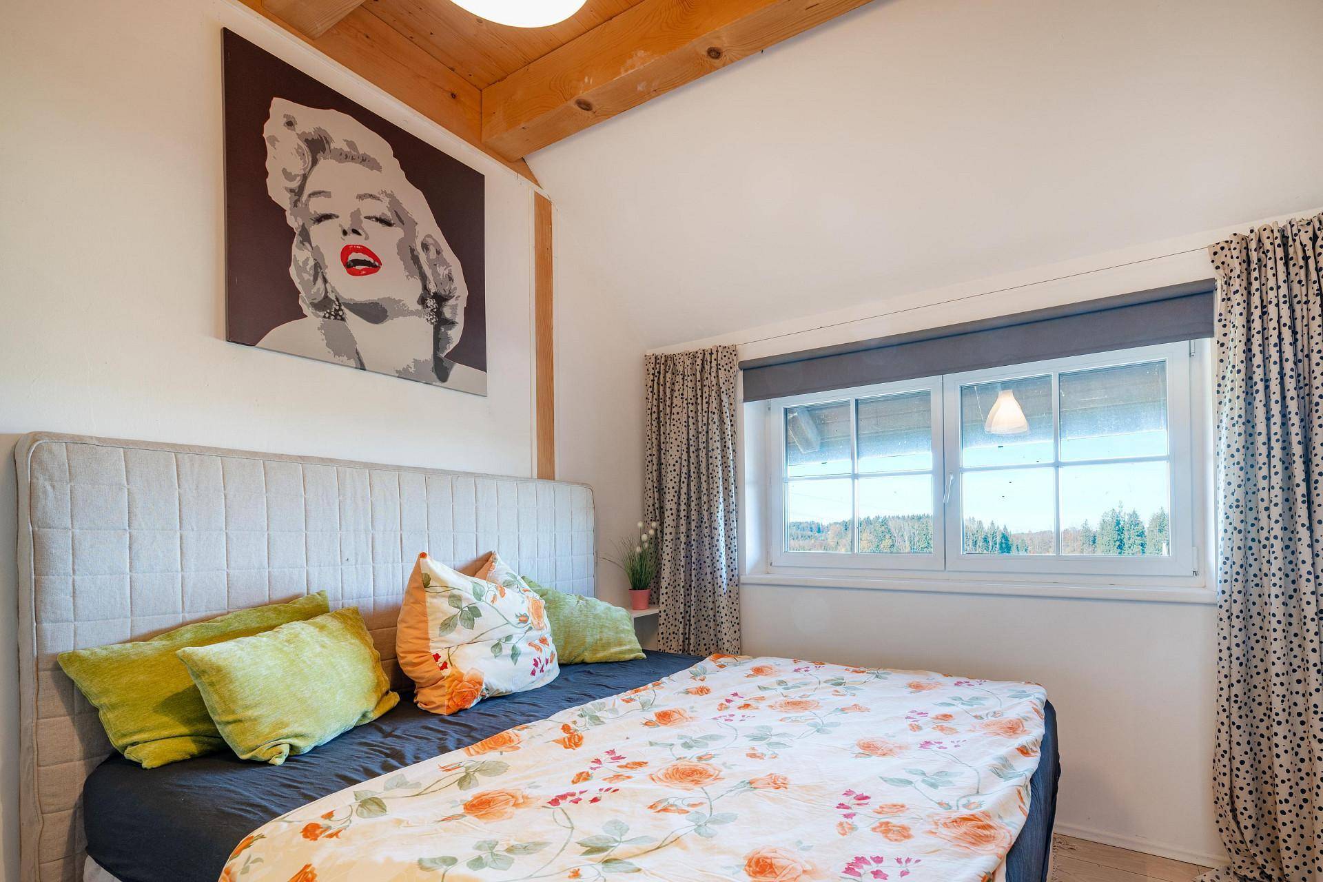 Helles Schlafzimmer mit Doppelbett, Holzbalken und Blick ins Grüne durch das Fenster.