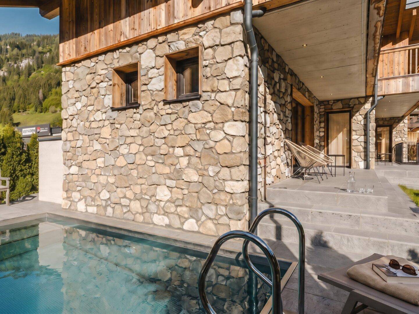 Außenbereich mit Natursteinfassade und kleinem Pool, umgeben von einer Terrasse mit Sitzgelegenheiten.