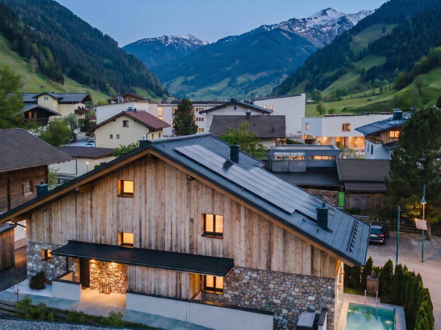 Abendansicht des Chalets mit Solaranlage auf dem Dach, Pool und beleuchteter Bergkulisse.