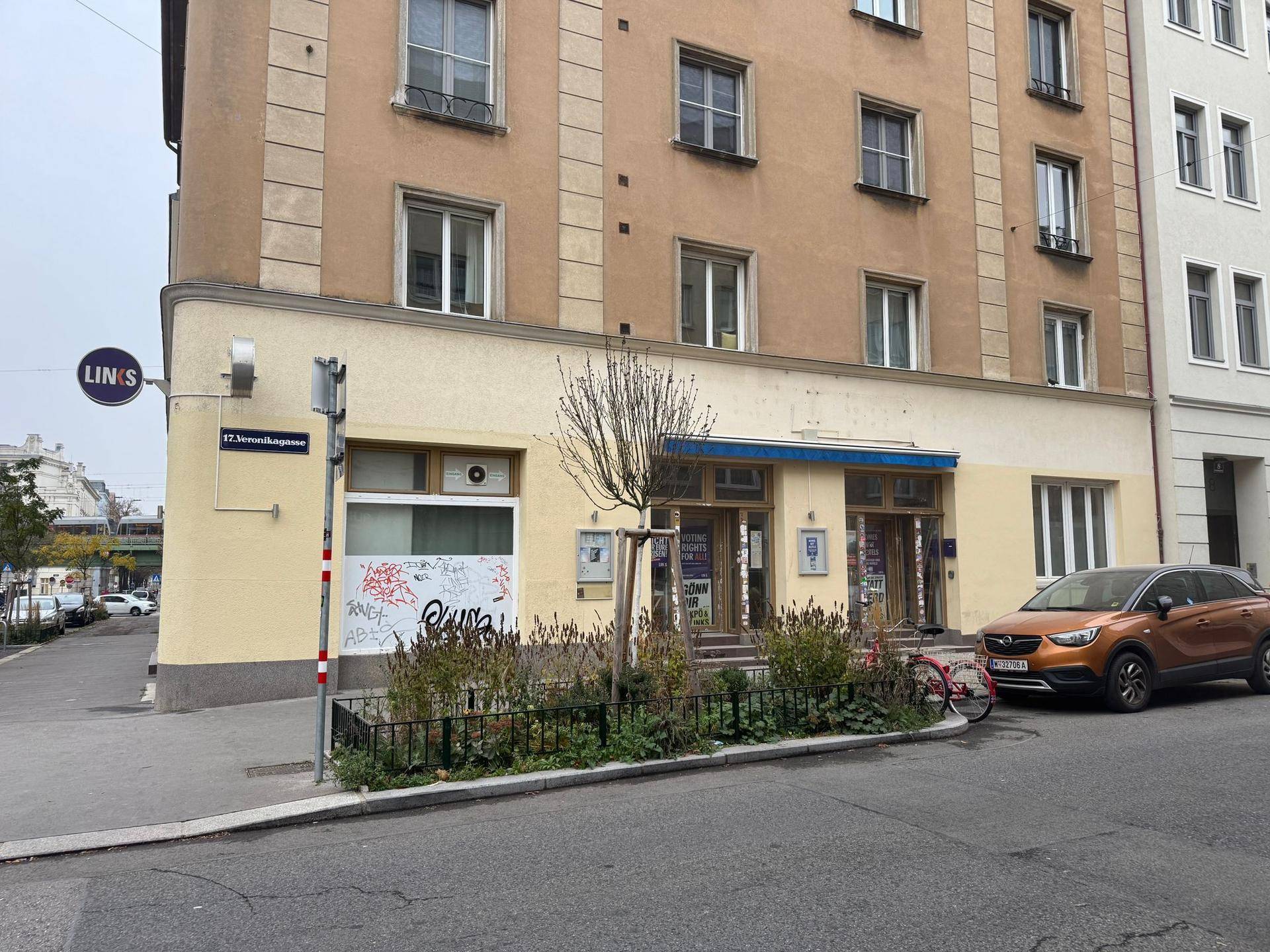 Gebäudeecke mit Ladenfront, Fenstern im Obergeschoss und einem geparkten Fahrzeug an der Straße.
