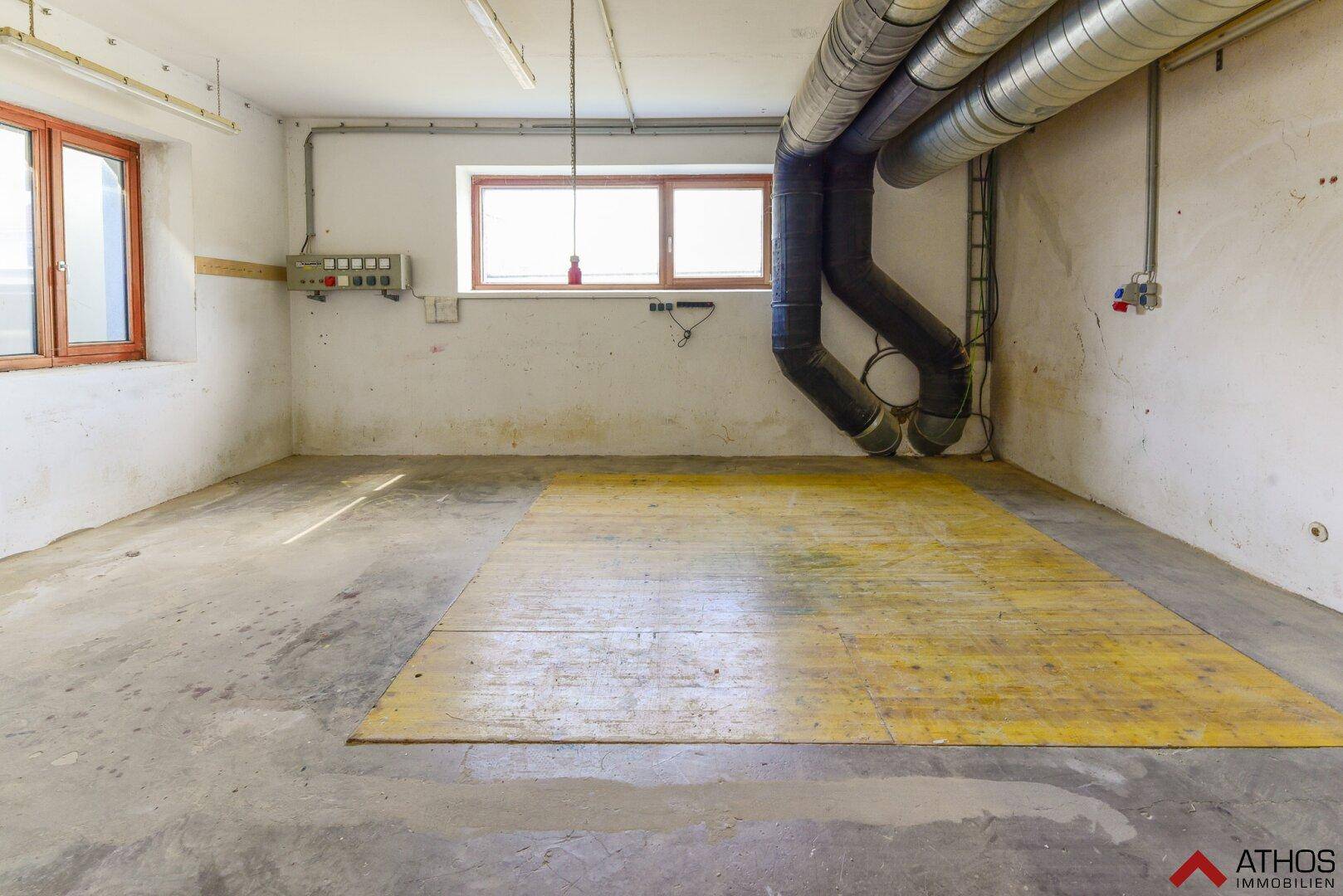 Einfacher Lagerraum mit Betonboden, kleinen Fenstern und großen Lüftungsrohren an der Wand.