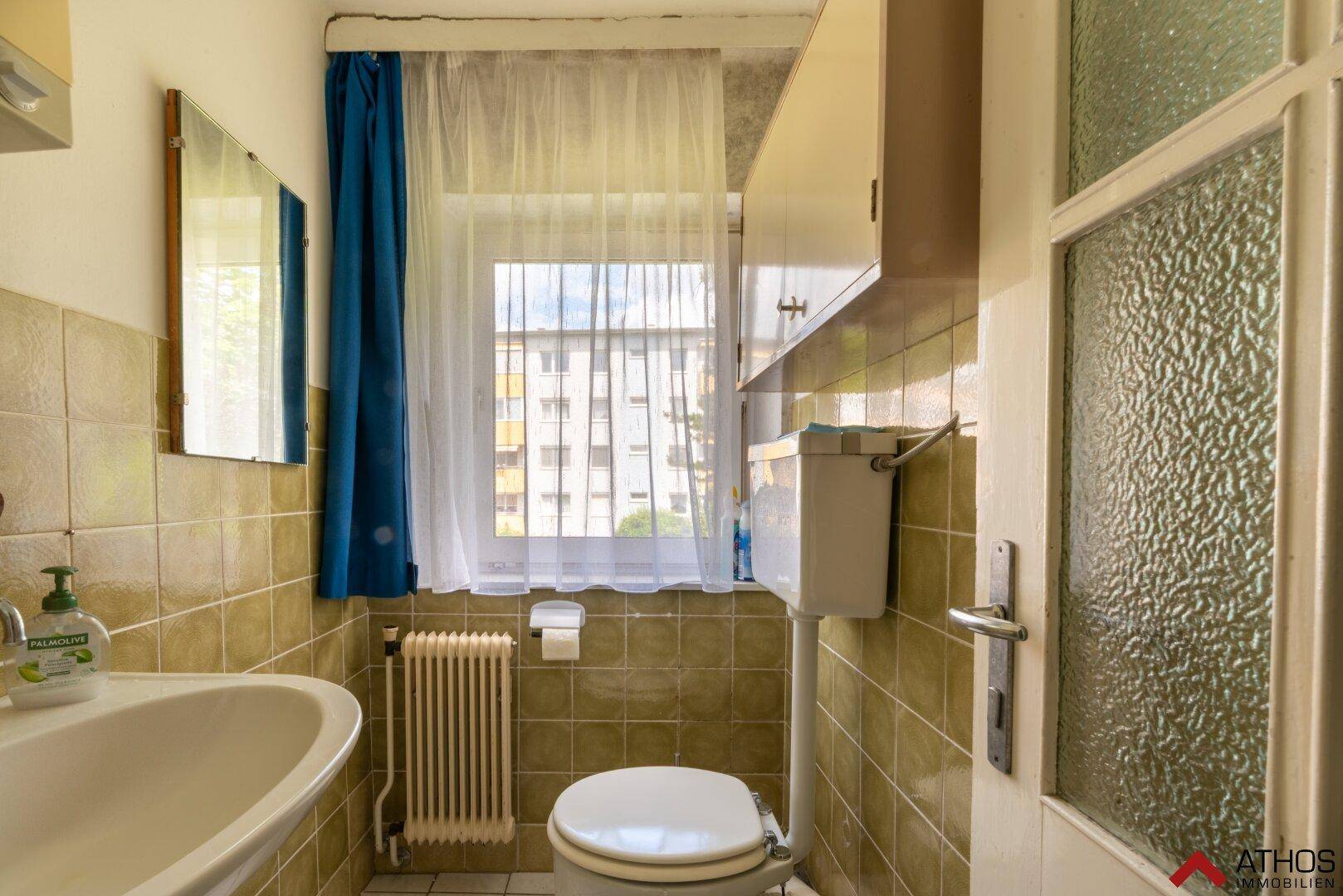 Kleines Badezimmer mit beigen Fliesen, WC, Waschbecken und einem Fenster für Tageslicht.