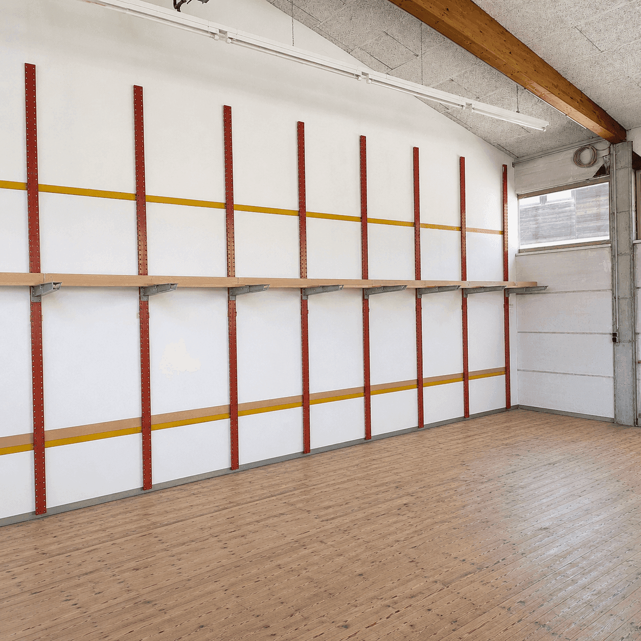 Heller Lagerraum mit Regalsystem an der Wand, Holzboden und Tageslicht durch ein Fenster.