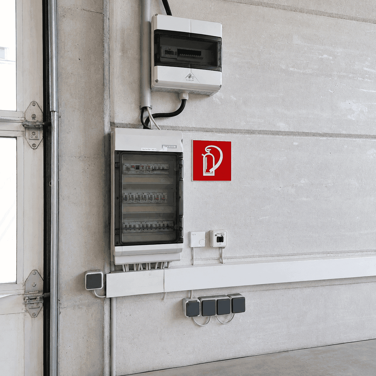 Betonwand mit elektrischen Schaltkästen, Steckdosen und einem Feuerlöscher-Hinweisschild.