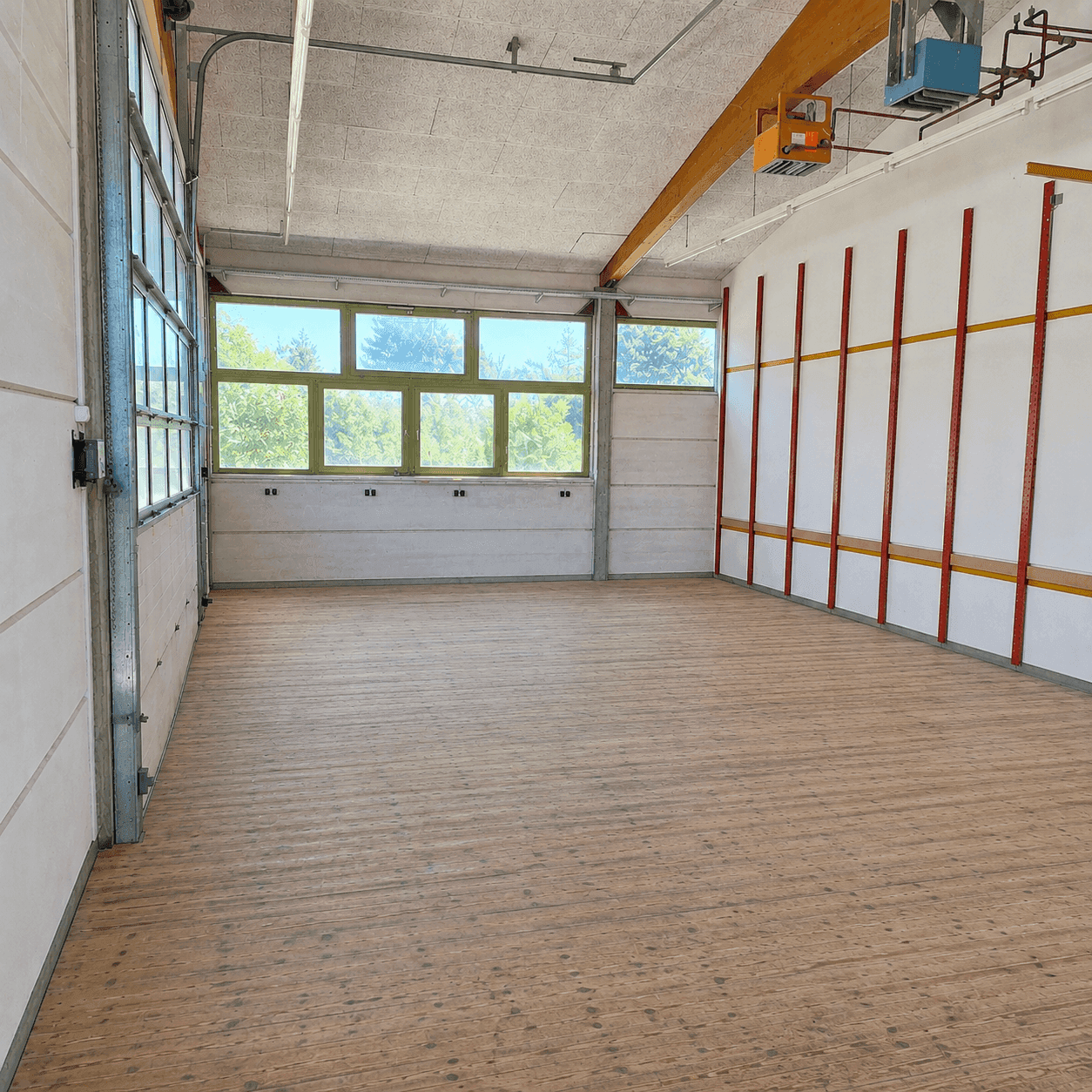 Geräumiger Innenraum mit Holzboden, großen Fenstern und sichtbaren Deckeninstallationen.