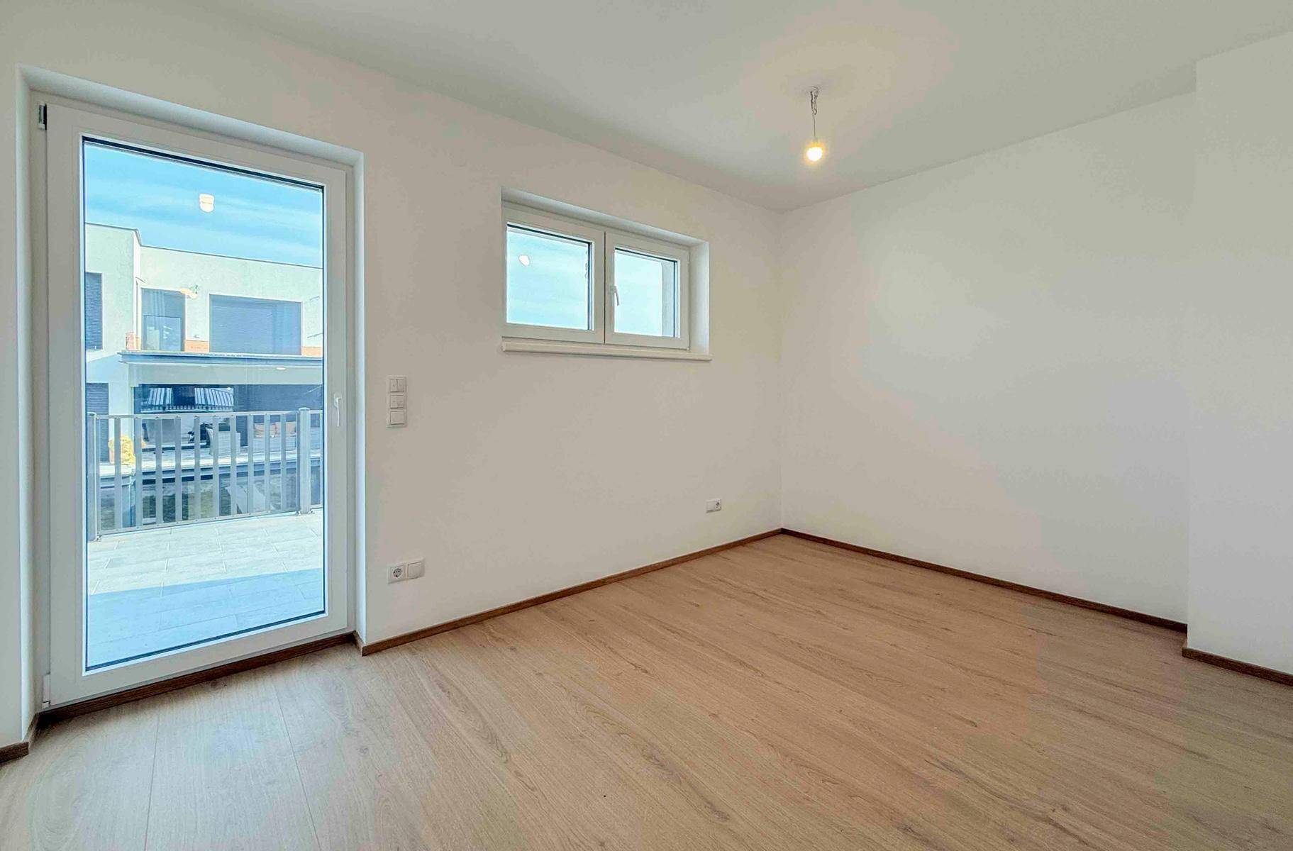 Leerer Raum mit Laminatboden und Zugang zu einem Balkon, bietet viel Tageslicht.
