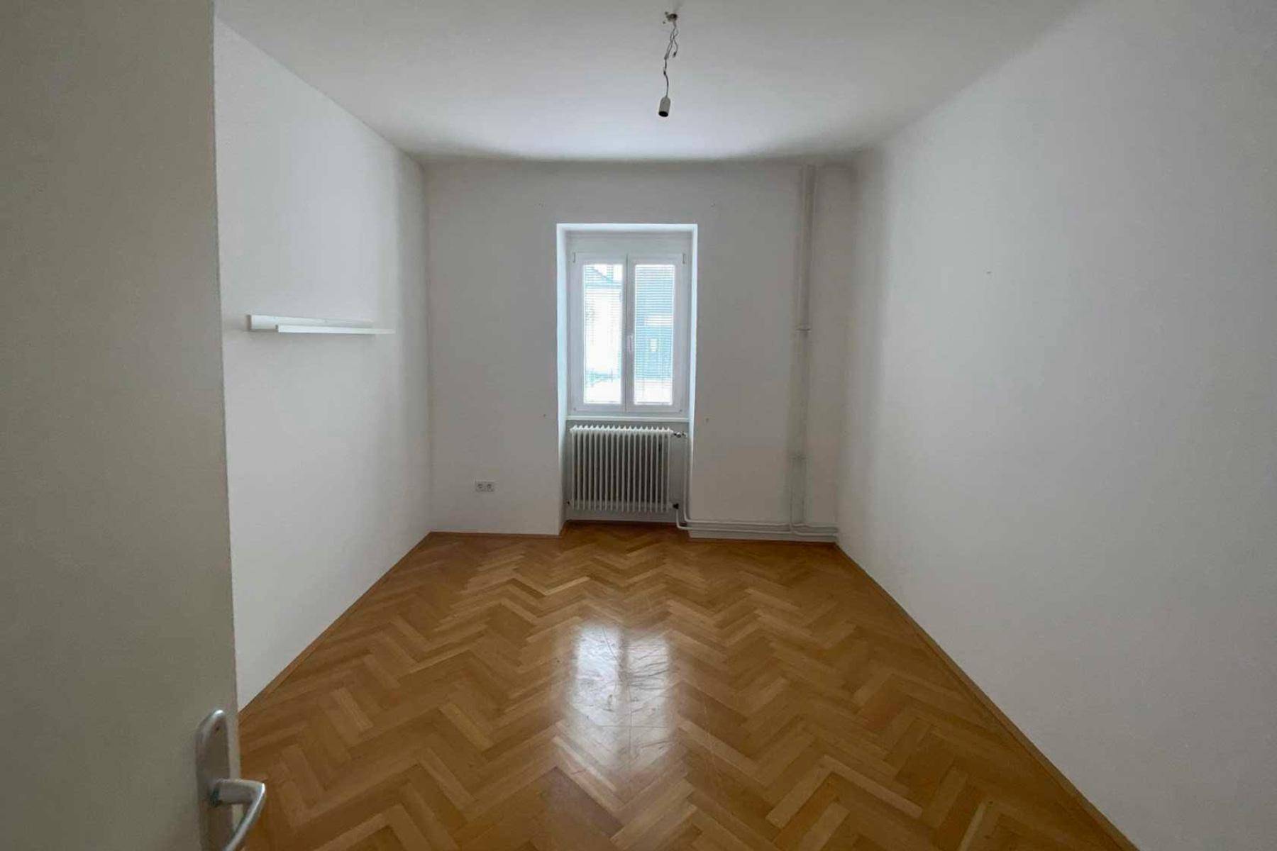 Helles Schlafzimmer mit Fischgrätparkett und einem Fenster, das viel Tageslicht hereinlässt.