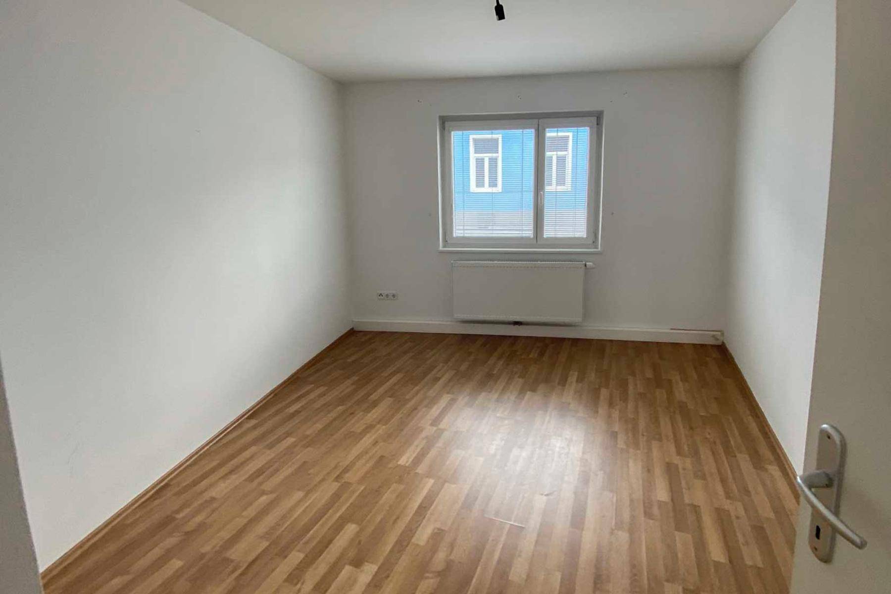 Einfaches Schlafzimmer mit Laminatboden und einem Fenster, das den Raum mit Tageslicht versorgt.