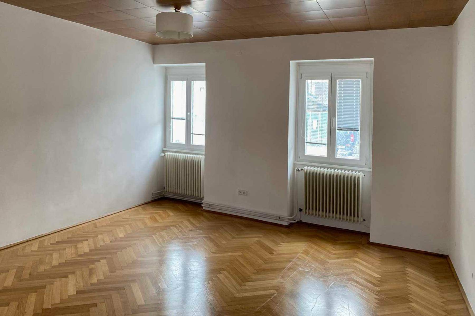 Heller Raum mit Fischgrätparkett, zwei Fenstern und einer Holzdecke, die eine gemütliche Atmosphäre schafft.