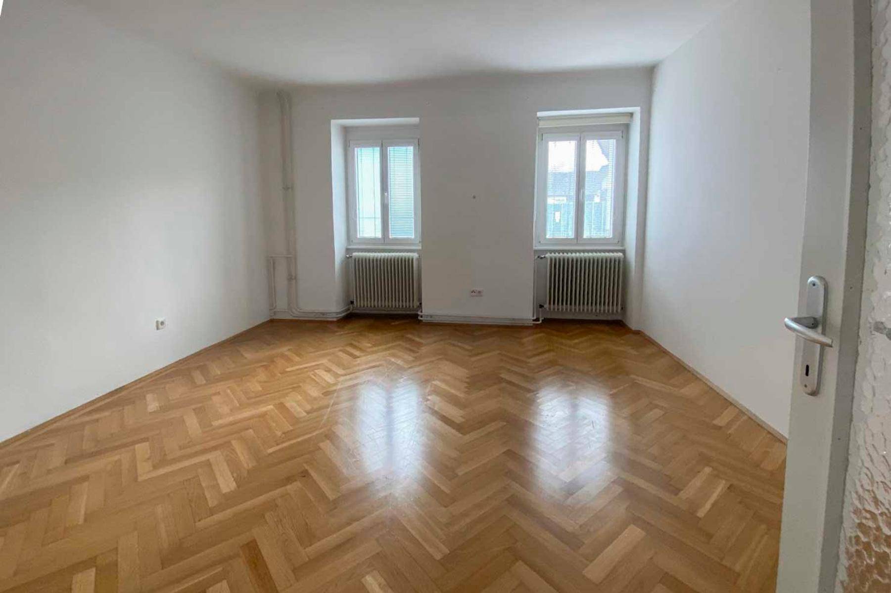 Geräumiges Schlafzimmer mit klassischem Fischgrätparkett und zwei Fenstern für gute Belichtung.
