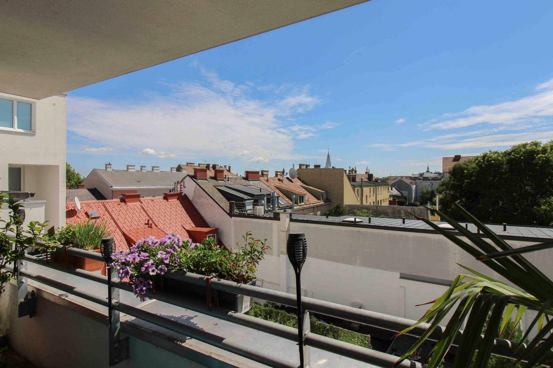Blick vom Balkon über die Dächer der Stadt mit blühenden Pflanzen und blauem Himmel.