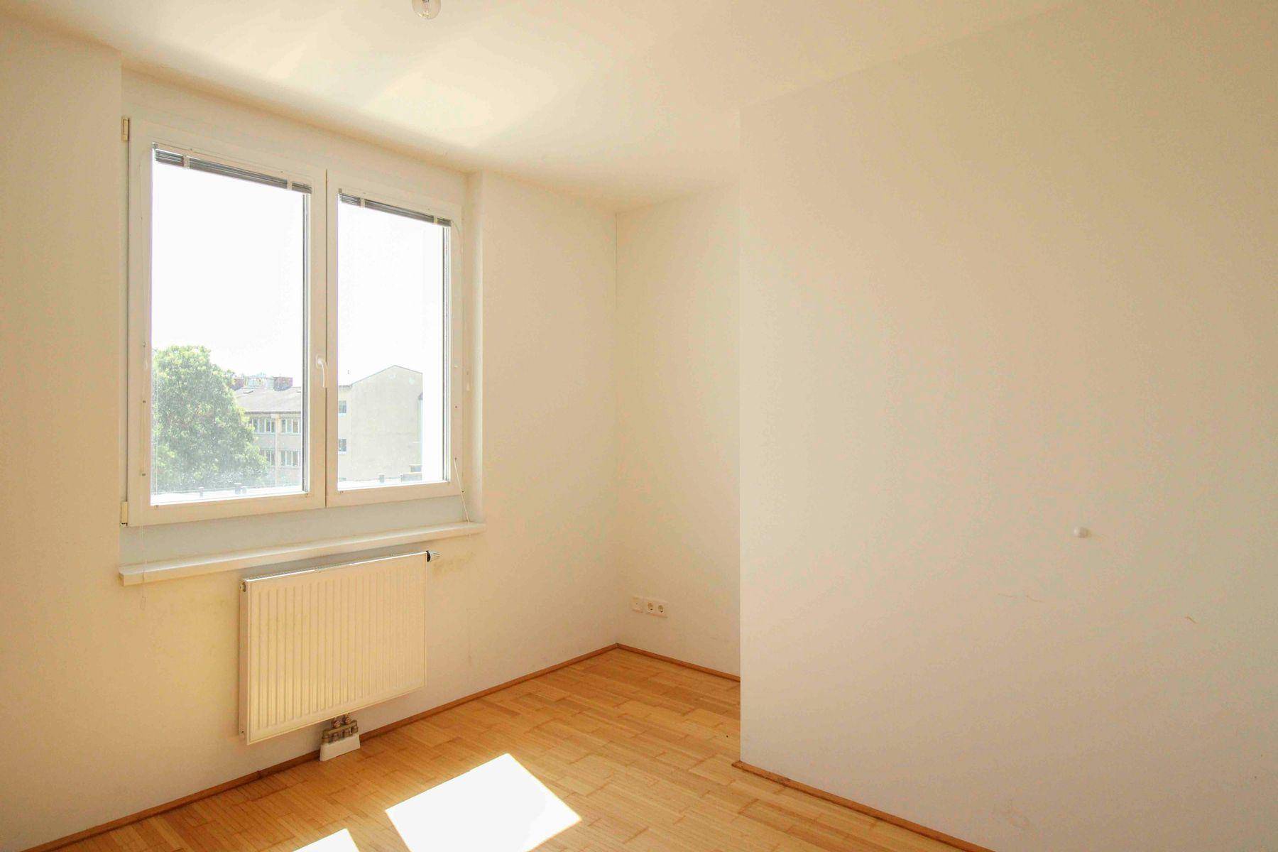 Leerer Raum mit hellem Parkettboden und großem Fenster, das viel Licht hereinlässt.