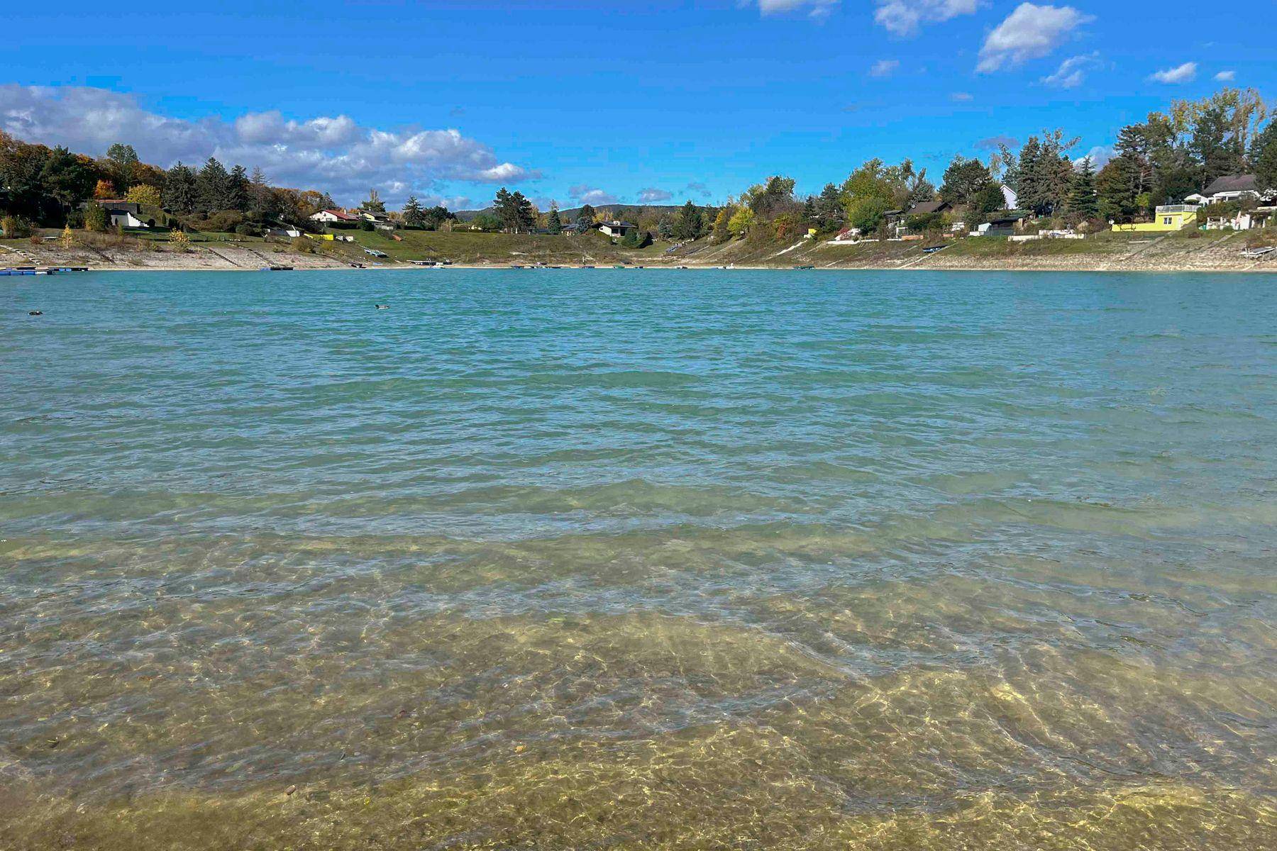 Klarer See mit ruhigem Wasser und bewaldeten Ufern unter blauem Himmel.