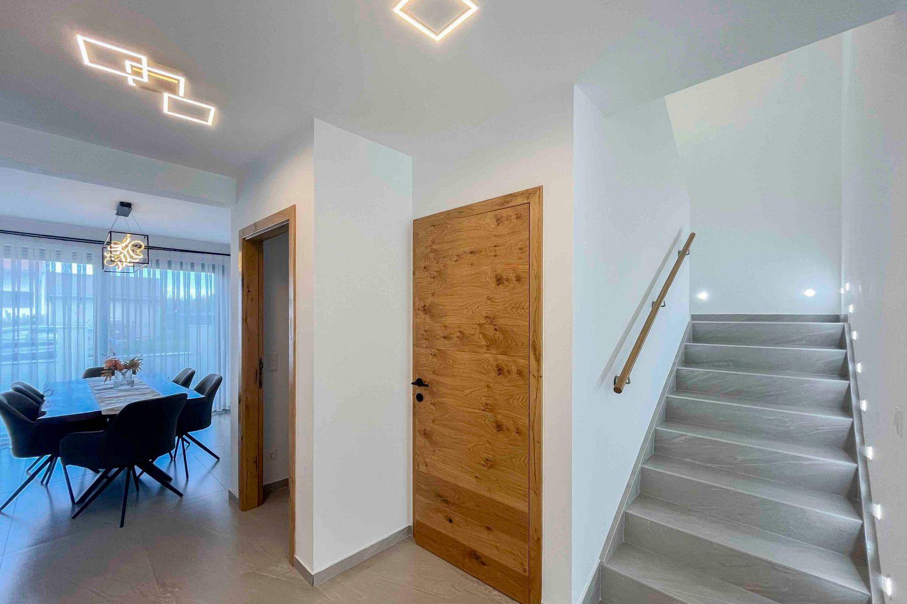 Moderner Treppenaufgang mit Fliesenstufen und Holzgeländer, beleuchtet durch Wandleuchten.