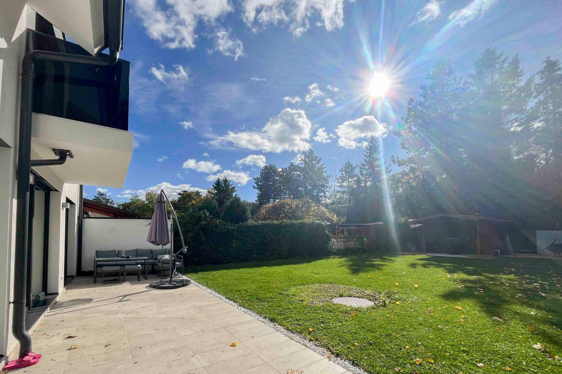 Sonnige Terrasse mit Gartenmöbeln und weitläufigem Rasenbereich, umgeben von Bäumen.