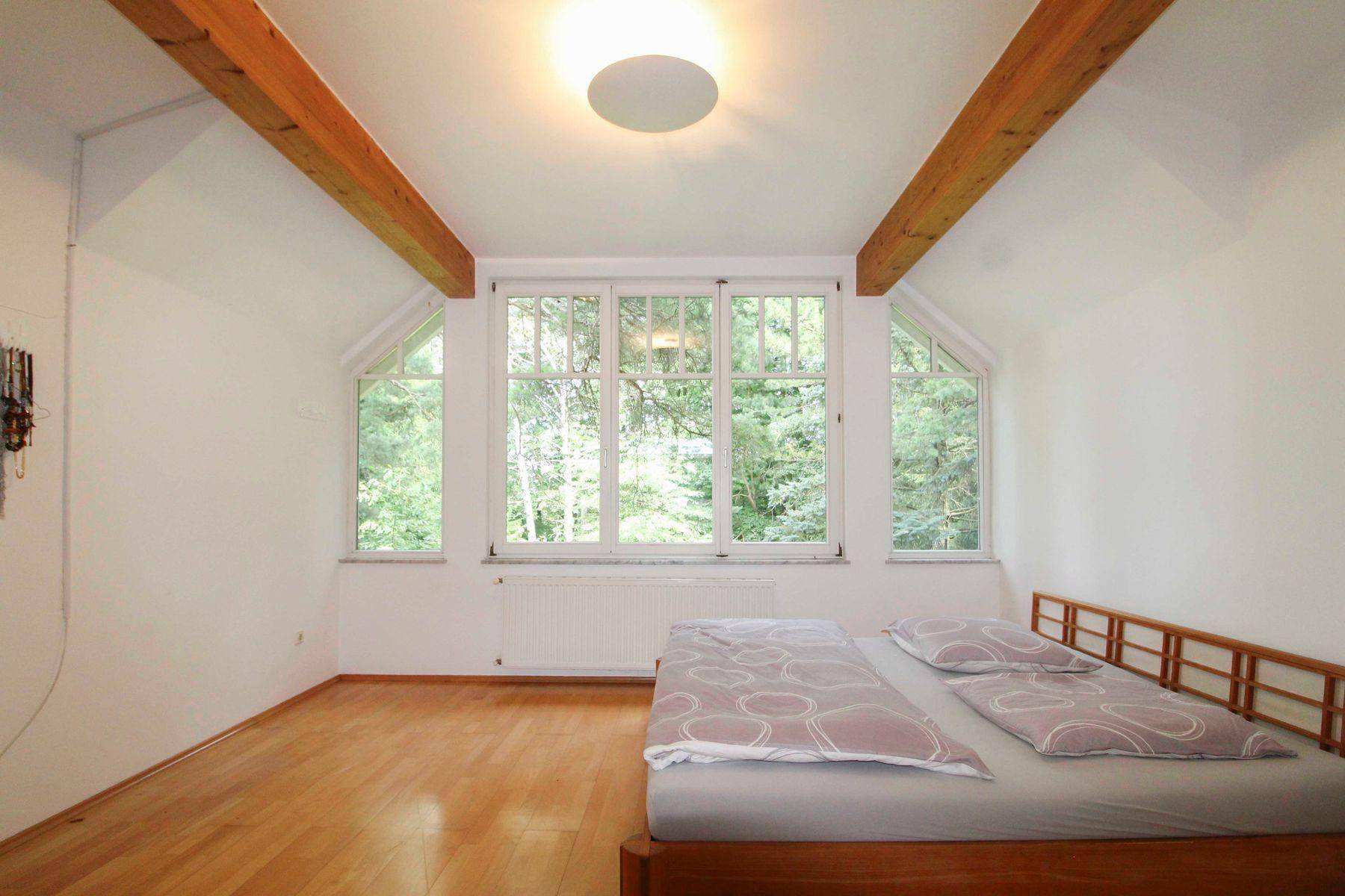 Schlafzimmer mit Holzboden, sichtbaren Holzbalken und großen Fenstern mit Blick ins Grüne.