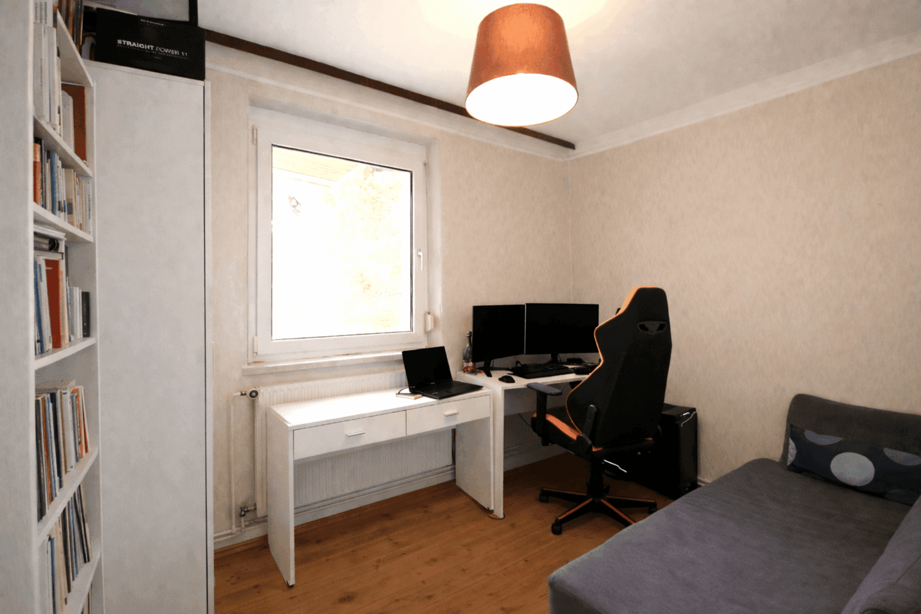 Helles Arbeitszimmer mit Holzfußboden, großem Fenster, Schreibtisch mit Monitoren und einem Bücherregal.