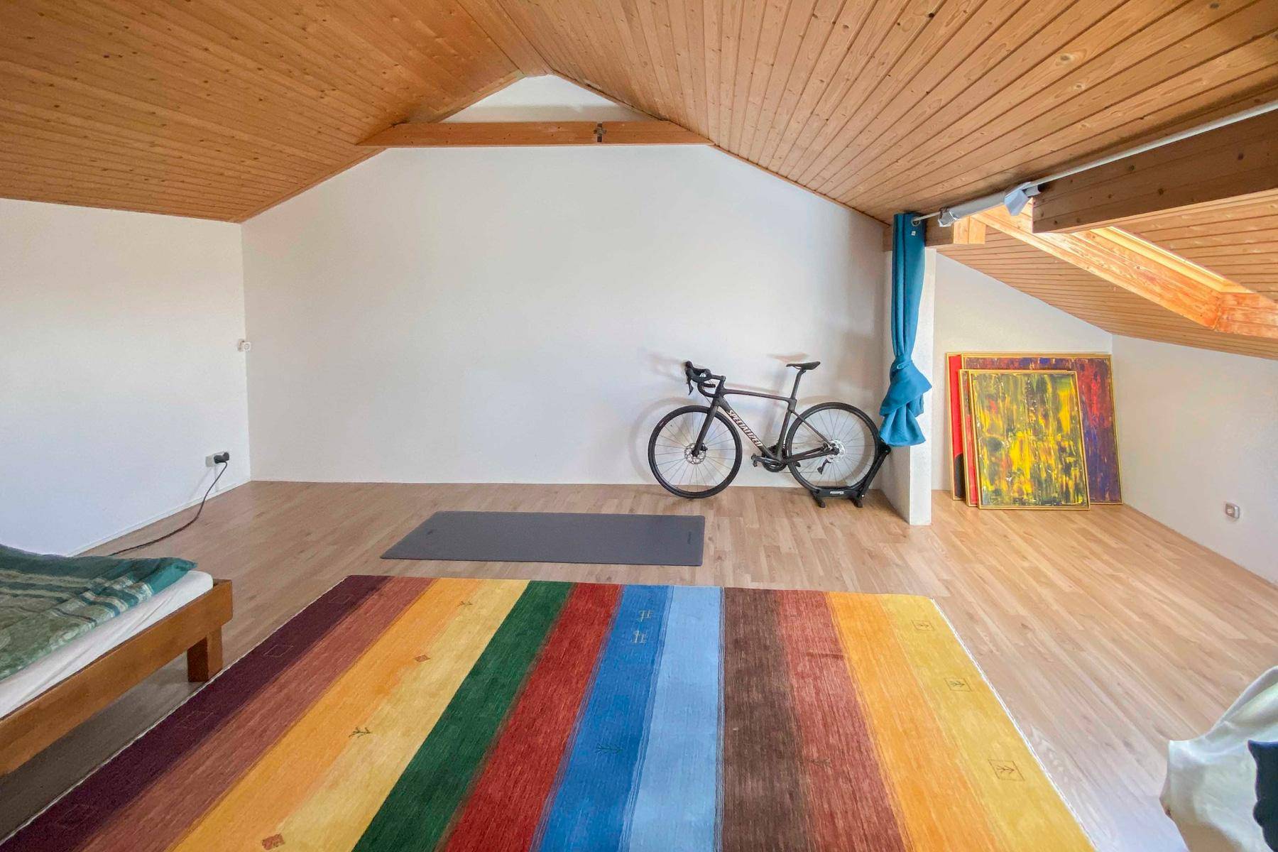 Großes Dachzimmer mit Holzbalkendecke und farbenfrohem Teppich, vielseitig nutzbar als Hobby- oder Fitnessraum.