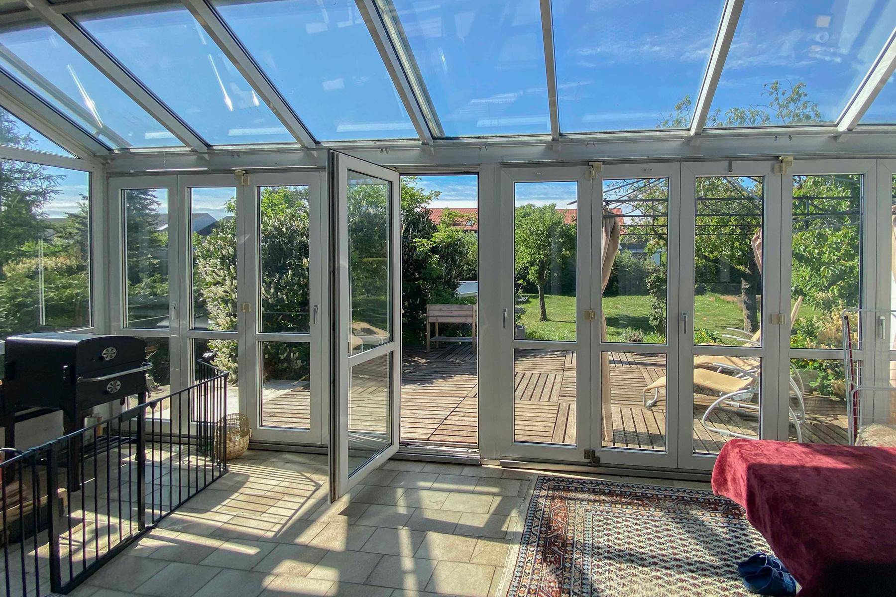 Heller Wintergarten mit Glasdach und Fliesenboden, bietet direkten Zugang zur Holzterrasse und zum Garten.