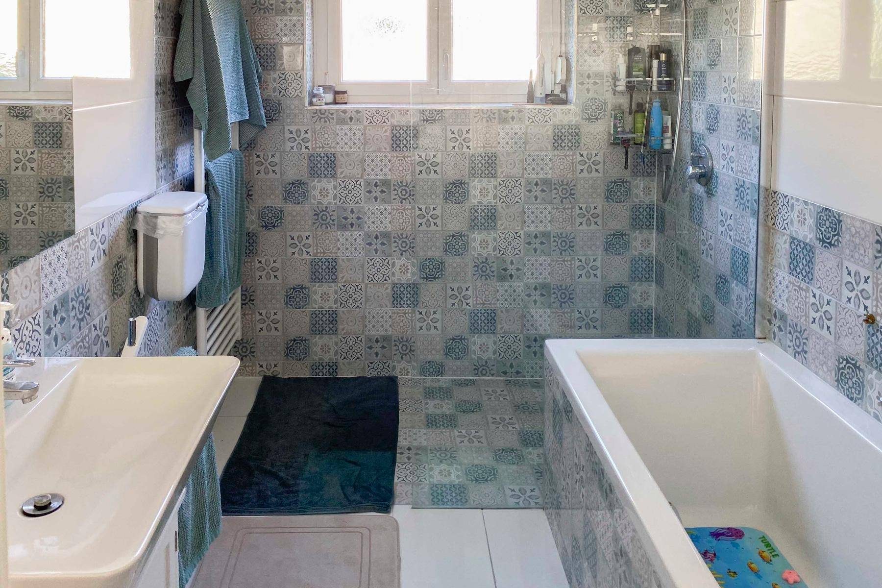 Badezimmer mit gemusterten blau-weißen Fliesen, Badewanne mit Duschfunktion und Fenster für Tageslicht.