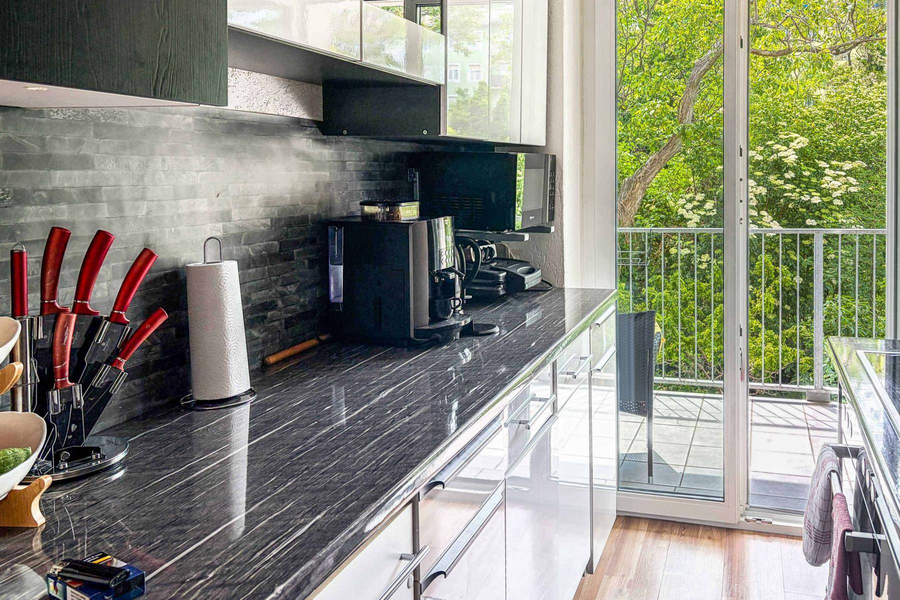 Moderne Küche mit dunkler Arbeitsplatte, glänzenden weißen Schränken und Zugang zu einem Balkon mit Grünblick.