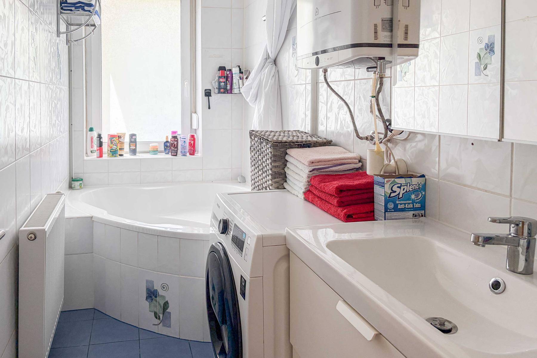 Helles Badezimmer mit Eckbadewanne, Waschbecken und integrierter Waschmaschine, alles in weiß gefliest.