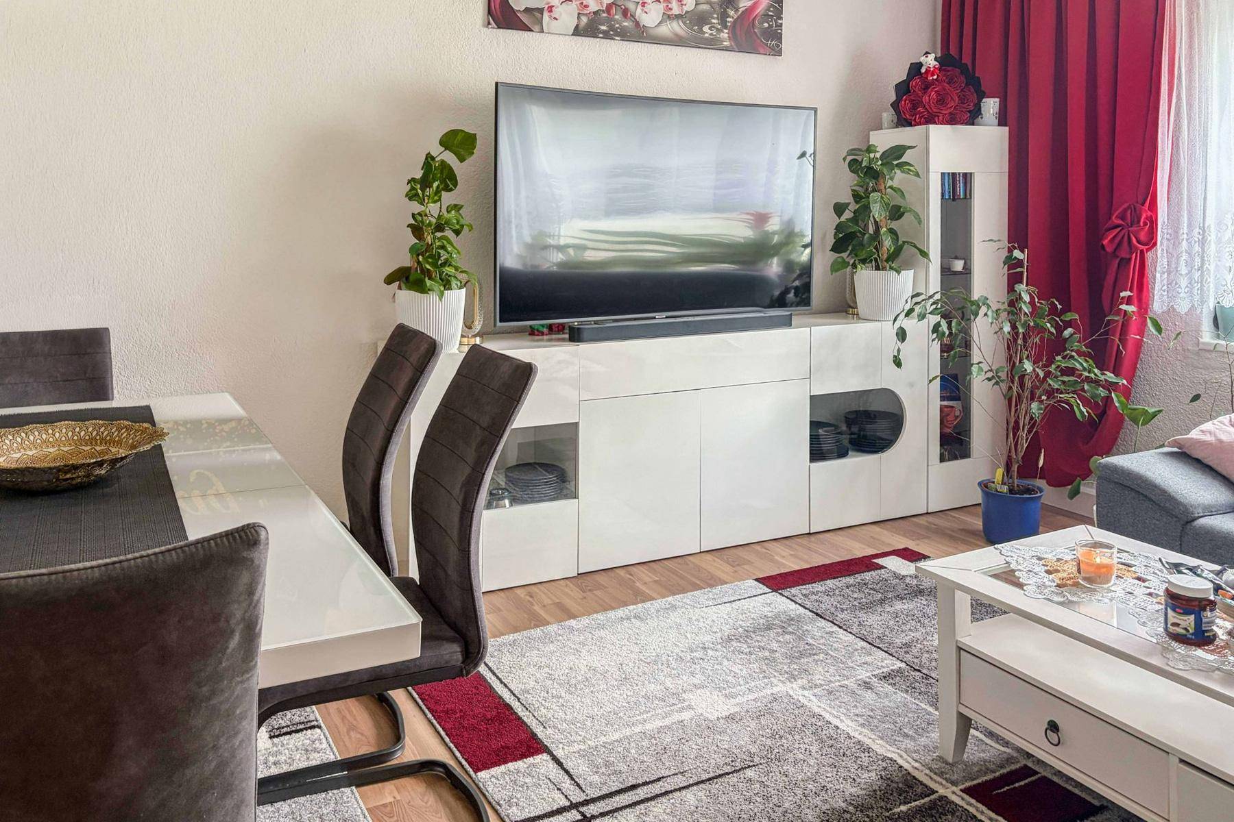Wohnbereich mit modernem TV-Möbel, Esstisch und Stühlen, ergänzt durch Pflanzen und rote Vorhänge.
