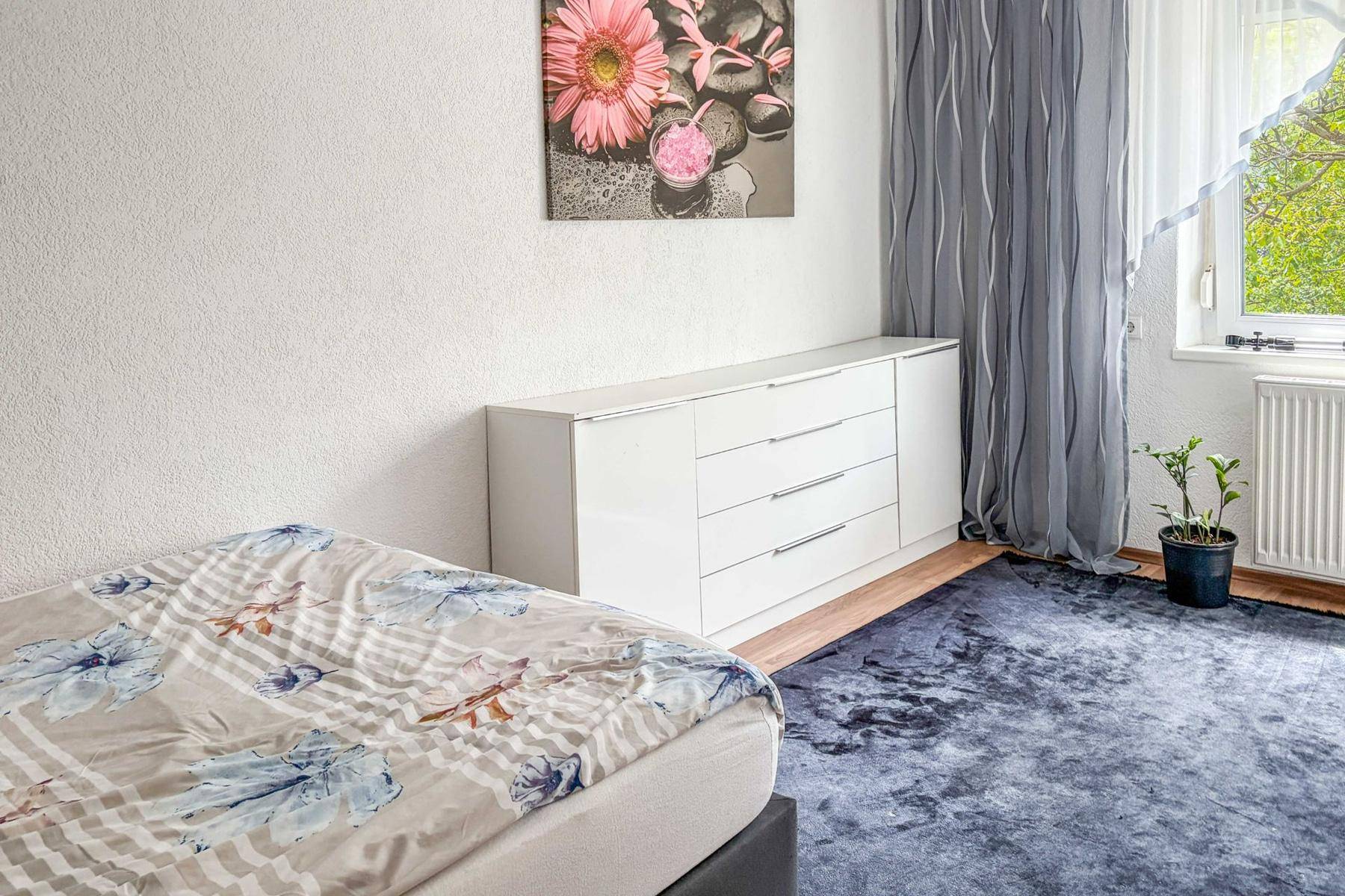 Schlafzimmer mit Einzelbett, weißer Kommode und einem Fenster, das für natürliche Beleuchtung sorgt.
