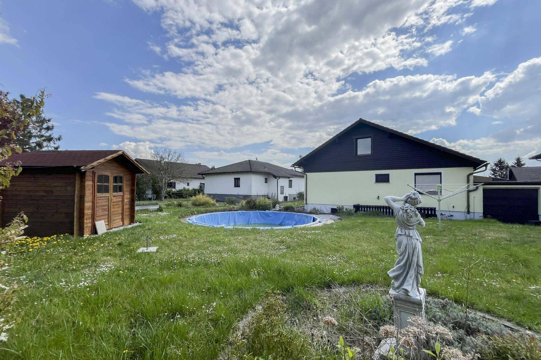 Hinterhof mit Gartenhaus, einem runden Pool und einer Gartenstatue unter weitem Himmel.