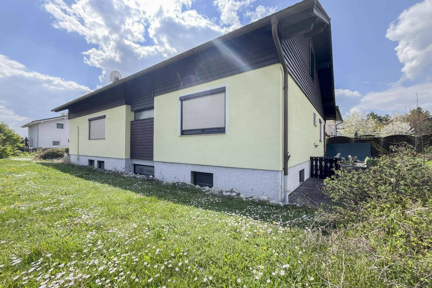 Seitenansicht des Hauses mit Garten, kleinen Fenstern und Rollläden unter sonnigem Himmel.