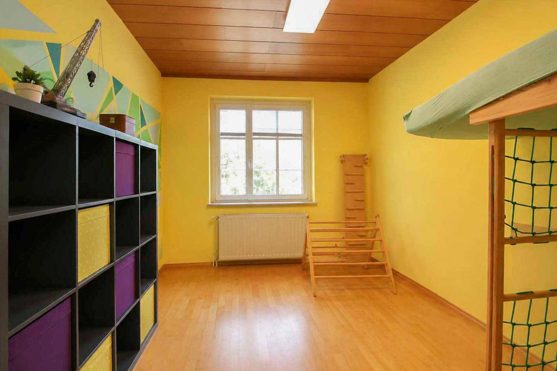 Helles Kinderzimmer mit gelben Wänden, Holzdecke und Spielgeräten.